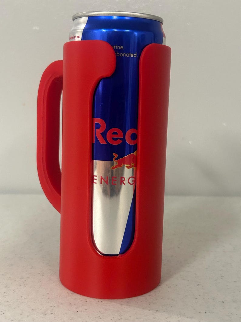 May include: A red can cooler with a handle holds a blue and silver Red Bull energy drink can. The cooler has a curved design to fit the can snugly. The Red Bull can has the word "Red" in large letters.