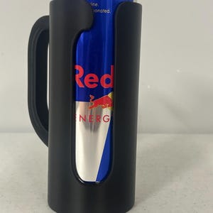 May include: A black drink holder with a handle, holding a blue and silver Red Bull energy drink can. The can has the word "Red" in red and a red bull logo. The words "Energy Drink" are also visible.