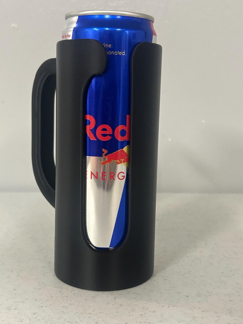 May include: A black drink holder with a handle, holding a blue and silver Red Bull energy drink can. The can has the word "Red" in red and a red bull logo. The words "Energy Drink" are also visible.