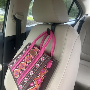 May include: A black plastic car headrest hook attached to a beige seat, holding a small brown handbag with pink handles and a vibrant geometric pattern in shades of orange, pink, and white, featuring a rectangular brown brand label on the front.