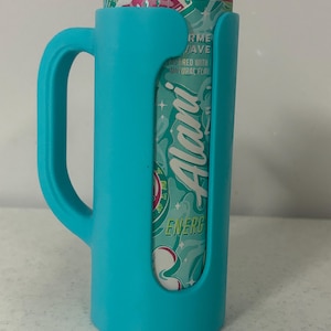 May include: A teal-colored can cooler with a handle, holding a can of Alani Energy drink. The can is teal and pink, with the Alani logo visible. The cooler has a handle for easy carrying.