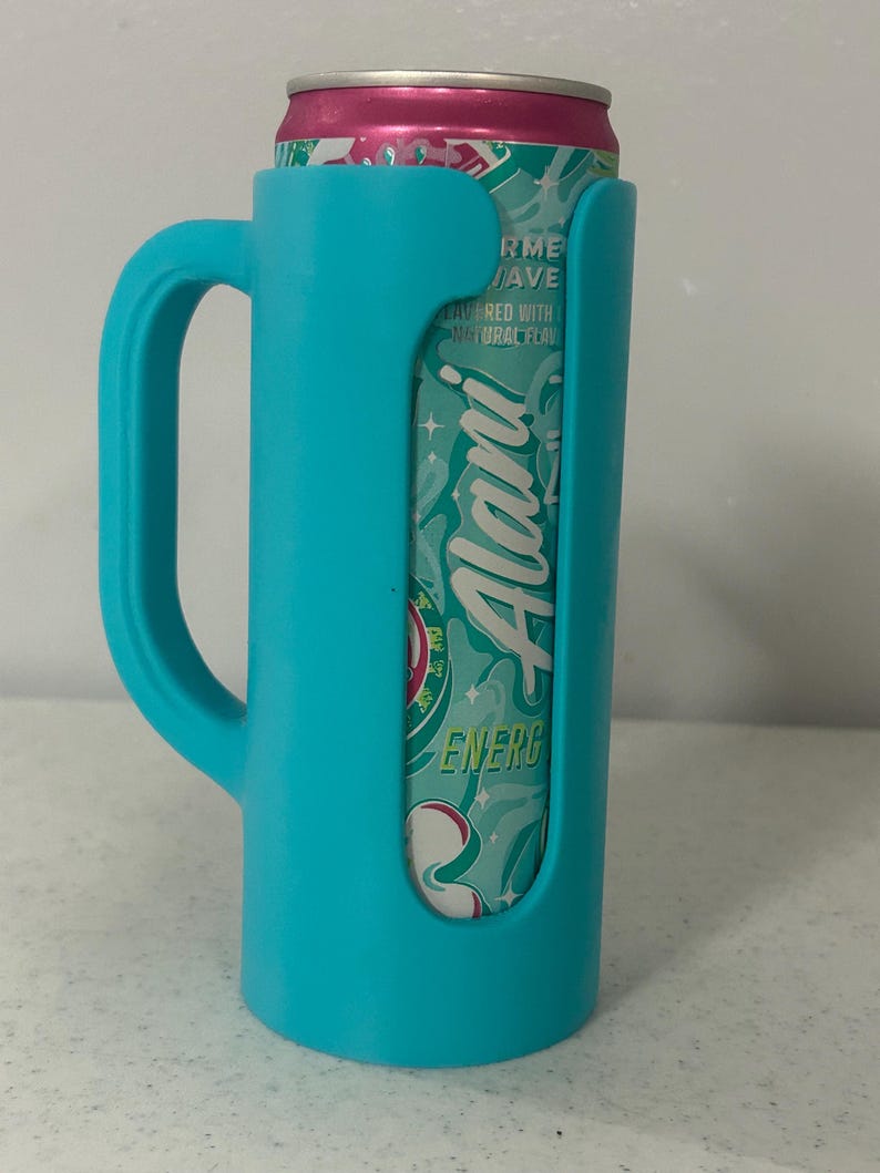 May include: A teal-colored can cooler with a handle, holding a can of Alani Energy drink. The can is teal and pink, with the Alani logo visible. The cooler has a handle for easy carrying.