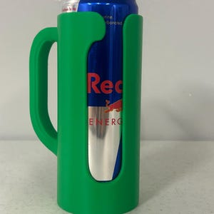 May include: A green plastic can holder with a handle, holding a Red Bull energy drink can. The can is blue with red and silver branding. The holder has a cutout to display the can's label.