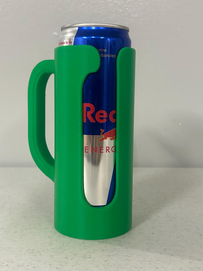 May include: A green plastic can holder with a handle, holding a Red Bull energy drink can. The can is blue with red and silver branding. The holder has a cutout to display the can's label.