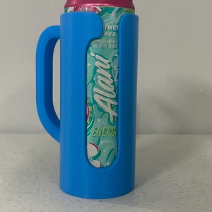 May include: A blue can cooler with a handle, holding a can of Alani Nu Energy drink. The can is pink and teal with the Alani Nu logo visible. The cooler is made of plastic and designed to keep beverages cold.