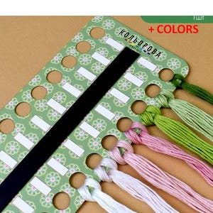 May include: A green and white thread organizer with a black strap and several colored thread bundles. The organizer has a floral pattern and the text "+ COLORS" is visible. The thread colors include white, pink, and green.