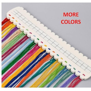 May include: A white thread organizer with a grid pattern and colorful embroidery floss. The floss includes shades of red, orange, yellow, green, blue, and purple. The text "MORE COLORS" is displayed in red.