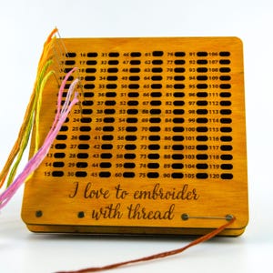 May include: A wooden embroidery thread organizer with numbered holes and colorful threads. The text "I love to embroider with thread" is printed on the front. A safety pin secures the thread.
