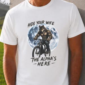 Può includere: T-shirt bianca con un lupo muscoloso in bicicletta davanti a una luna piena. Le scritte "HIDE YOUR WIFE" e "THE ALPHA'S HERE" sono stampate sulla maglietta. La parola "WHITE" è stampata in alto.
