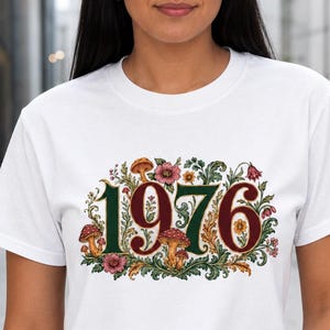 May include: A white short-sleeved t-shirt featuring a graphic print of the number 1976 in a dark red font. The numbers are surrounded by detailed illustrations of green foliage, various colorful flowers, and small brown mushrooms.