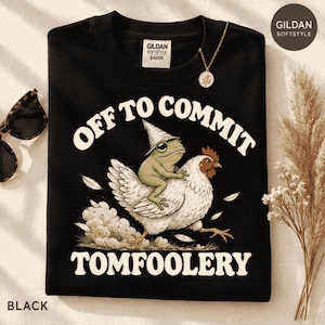 May include: A black t-shirt featuring a graphic of a green frog wearing a pointed hat riding a white chicken. The text above and below the illustration reads OFF TO COMMIT TOMFOOLERY in a white, curved, bold, retro-style font. The shirt has a Gildan Softstyle label.