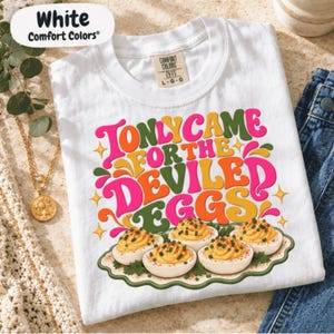May include: A white short-sleeve t-shirt featuring the colorful text 'I ONLY CAME FOR THE DEVILED EGGS' in a retro font, accompanied by a graphic illustration of four deviled eggs on a scalloped platter with green garnish.