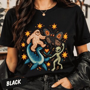 May include: A black t-shirt featuring a colorful graphic print of a white Bigfoot, a moth, a blue aquatic creature, and a green alien dancing together. The design is surrounded by yellow stars and a small heart, with the word BLACK printed in white.