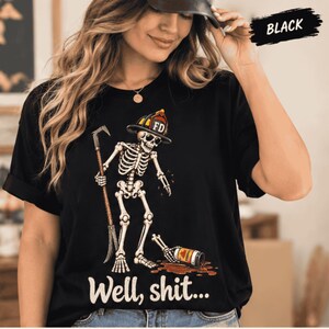 May include: A black t-shirt featuring a graphic of a skeleton wearing a firefighter helmet and sunglasses, holding a tool, standing over a spilled bottle with a puddle. The text "Well, shit..." is printed below the graphic, and "BLACK" is at the top.