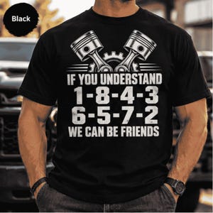 May include: A black short-sleeve t-shirt featuring a white graphic of two crossed engine pistons and a gear. The text reads: IF YOU UNDERSTAND 1-8-4-3 6-5-7-2 WE CAN BE FRIENDS. The design is centered on the chest of the garment.