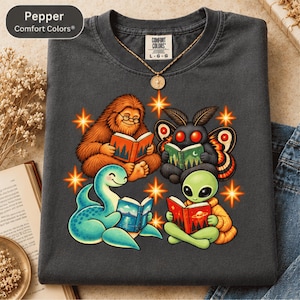 May include: A charcoal gray t-shirt featuring a graphic of a brown Bigfoot, a black moth creature, a blue sea monster, and a green alien, all reading books. The shirt has a Comfort Colors label and a gold pendant necklace resting on the collar.