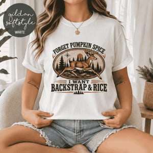 May include: A white short-sleeve t-shirt featuring a graphic of a running deer in a forest landscape. The text reads FORGET PUMPKIN SPICE I WANT BACKSTRAP & RICE. The design includes a compass and pine trees in brown and black tones.