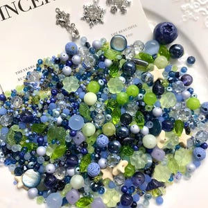 Op de afbeelding: Een verzameling kralen en bedels in blauwe, groene en witte tinten, waaronder sneeuwvlokbedels en diverse kraalvormen. De tekst "INCERE TO" is zichtbaar op de achtergrond. De kralen zijn gerangschikt op een wit bord.