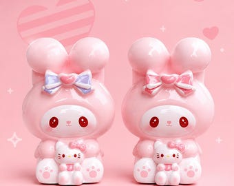 Puede incluir: Dos figuras rosas con forma de conejito con grandes orejas redondas y lazos. Cada figura tiene una pequeña figura de Hello Kitty en sus brazos. El fondo es de color rosa liso con formas de corazón y lazo.