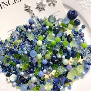 Könnte beinhalten: Eine Sammlung verschiedener Bastelperlen in Blau-, Grün- und Cremetönen mit verschiedenen Formen wie Kugeln, Sternen, Blättern und Blumen. Drei silberfarbene Metallanhänger in Schneeflocken- und Tropfenform liegen auf einer weißen Karte.