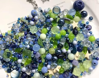 May include: A collection of assorted craft beads in shades of blue, green, and cream, featuring various shapes including spheres, stars, leaves, and flowers. Three silver-toned metal snowflake and teardrop charms are placed on a white card.