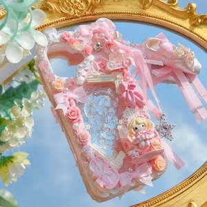Puede incluir: Una funda de teléfono rosa decorada con rosas, lazos y perlas. La funda presenta una muñeca en miniatura, zapatillas de ballet y un violín. El diseño incluye una sección transparente con un fondo azul, creando una estética caprichosa y femenina.