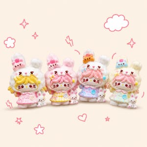 May include: Four colourful figurines of bunny-like characters with large ears, each wearing a dress and a hat with a small dessert on top. The figurines are in shades of pink, yellow, and blue, with white fur-like details. Small bunny figures are at their feet.