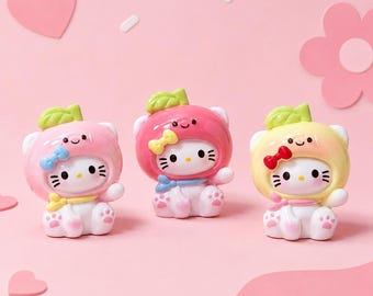 Puede incluir: Tres figuras de Hello Kitty en rosa, rojo y amarillo, cada una con un sombrero con temática de frutas con un lazo y una hoja verde. Las figuras tienen ojos negros, bigotes y huellas de patas en los pies. El fondo es de color rosa claro.