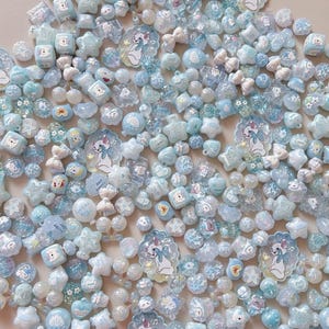 Op de afbeelding: Een grote verzameling diverse lichtblauwe en witte knutselkralen in verschillende vormen, waaronder sterren, harten, strikjes en ronde bollen, met diverse platte bedels die een wit katje met een blauwe strik afbeelden.