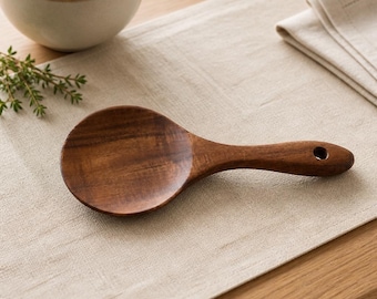May include: A dark brown wooden serving spoon with a wide, rounded bowl and a short, thick handle featuring a small circular hole at the end for hanging. The spoon has a smooth, polished finish with visible wood grain patterns throughout the surface.