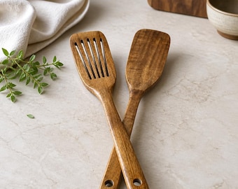 May include: Two dark brown wooden kitchen utensils, a slotted turner and a solid spatula, are crossed. Each handle features a circular hole at the end for hanging. The wood grain is visible throughout the surface of both cooking tools.