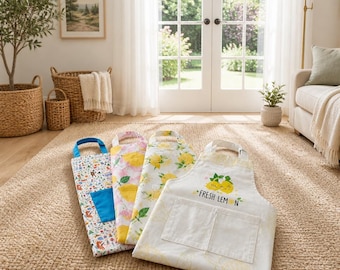 May include: Four aprons laid out on a rug. One features a colourful forest pattern with a blue pocket, one has a pink and yellow lemon print, one has a yellow lemon pattern, and one is white with a lemon graphic and the text FRESH LEMON.