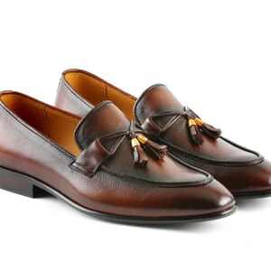 May include: A pair of brown leather loafers featuring a burnished finish, a rounded toe, and a decorative strap across the vamp. Each shoe is adorned with a leather tassel detail accented by small gold-toned metal hardware at the base.