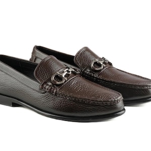 May include: A pair of brown leather loafers with a textured finish. Each shoe features a silver-toned metal ornament on the vamp and a black sole. The shoes are designed for a classic, sophisticated look.