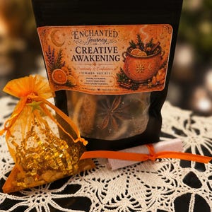May include: A black pouch labeled Enchanted Journey Creative Awakening Simmer Pot Kit with an orange organza bag filled with gold foil shreds and a small rolled paper tied with an orange ribbon, featuring illustrations of a cauldron and citrus.