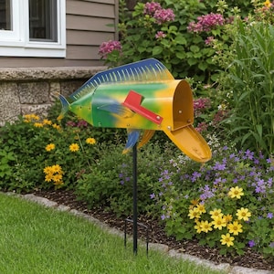 Könnte beinhalten: Eine dekorative, fischförmige Briefkasten in Gelb, Grün und Blau mit roter Fahne. Sie ist auf einem schwarzen Metallpfosten montiert und steht in einem Blumengarten.