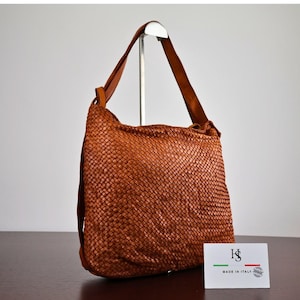 May include: A cognac-coloured woven leather hobo bag with a single shoulder strap. A small white rectangular card is placed in front, featuring the black logo HS, the text MADE IN ITALY, and a small graphic of the Italian flag in green, white, and red.