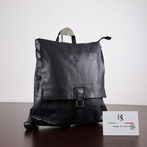 Peut inclure: Un sac à dos en cuir noir avec un rabat, une boucle couleur laiton et une poignée supérieure. Une carte rectangulaire blanche est placée devant, avec les lettres noires HS, le texte MADE IN ITALY et un petit drapeau italien.
