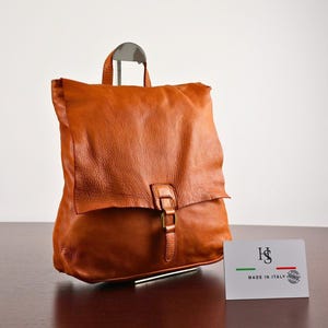 Op de afbeelding: Een cognac-kleurige leren rugzak met een handvat aan de bovenkant en een overslagflap die wordt vastgezet met een metalen gespriem. Voor de tas staat een wit rechthoekig kaartje met de letters HS, de kleuren van de Italiaanse vlag en MADE IN ITALY.
