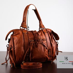 Op de afbeelding: Een cognac-kleurige leren handtas met twee handvatten, een voorvak met rits en kwastjes, en decoratieve metalen studs. Een klein wit kaartje naast de tas toont het logo HS en de tekst MADE IN ITALY.