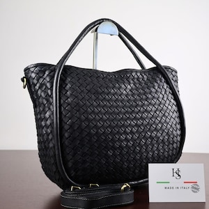 Può includere: Una borsa in pelle nera con motivo intrecciato, due manici arrotondati e una tracolla rimovibile con finiture color ottone. È presente un cartellino bianco con la scritta HS MADE IN ITALY e un piccolo grafico nei colori verde, bianco e rosso.