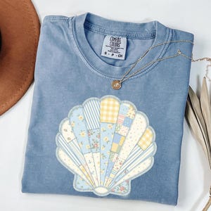 May include: A blue short-sleeved t-shirt featuring a large seashell graphic composed of various patchwork patterns in yellow, blue, and white floral, gingham, and anchor prints. The inner neck label reads Comfort Colors, S-P-CH, 100% cotton.