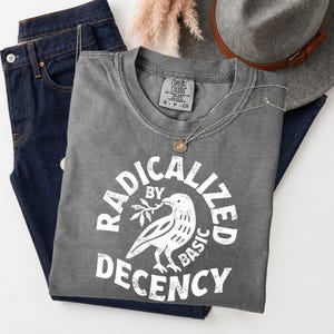 以下が含まれることがあります： 枝をくわえた鳥の白いグラフィックと「RADICALIZED BY DECENCY」という文字がプリントされたチャコールグレーのTシャツ。ダークブルーのデニムジーンズ、茶色のバンドが付いたグレーのフェルトハット、ゴールドのペンダントネックレスが添えられています。