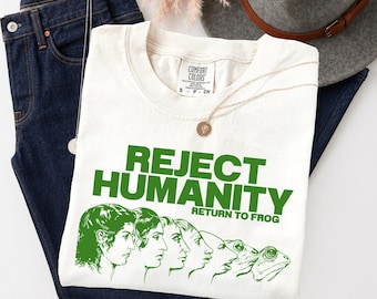 May include: A cream-colored short-sleeve t-shirt featuring green text that reads REJECT HUMANITY RETURN TO FROG. Below the text is a green graphic showing the evolutionary transition from a human profile to a frog profile in a series of stages.