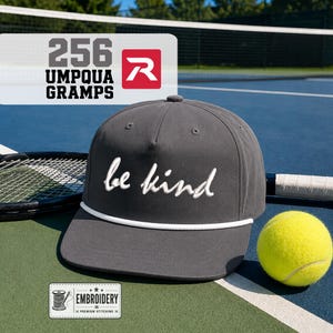May include: A charcoal gray baseball cap featuring the white embroidered text be kind in a cursive script. The cap includes a white rope detail across the base of the crown and a structured five-panel design with matching gray eyelets.