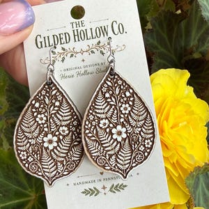May include: A pair of teardrop-shaped wooden earrings featuring a laser-engraved botanical pattern of white flowers and fern fronds. The earrings are attached to a display card labeled The Gilded Hollow Co. with silver-toned metal hooks.