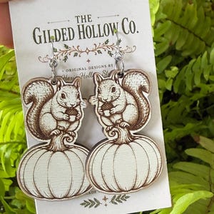 May include: A pair of dangle earrings featuring a brown and white illustration of a squirrel holding an acorn while sitting on a pumpkin. The earrings are attached to a white display card with the text The Gilded Hollow Co. printed on it.