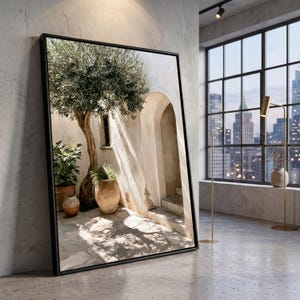 May include: A large rectangular framed wall art piece featuring a Mediterranean courtyard scene with an olive tree, terracotta pots, and an arched doorway. The image displays light and shadow patterns on stone flooring and a textured white wall.