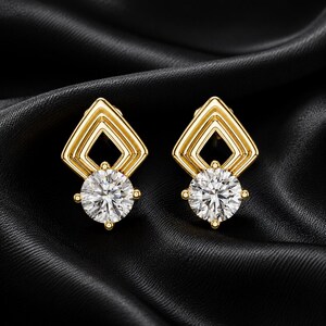 May include: A pair of gold-toned stud earrings featuring a geometric diamond-shaped frame at the top and a single round, clear, faceted stone set at the base. The earrings have a polished metallic finish and a secure prong setting for the stone.