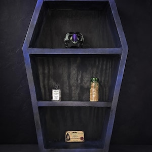 May include: A coffin-shaped wooden wall shelf with a dark purple and black finish. The shelf features three tiers holding a small black skull, a clear glass bottle with a white label, a glass jar with tan contents, and a rectangular death certificate.